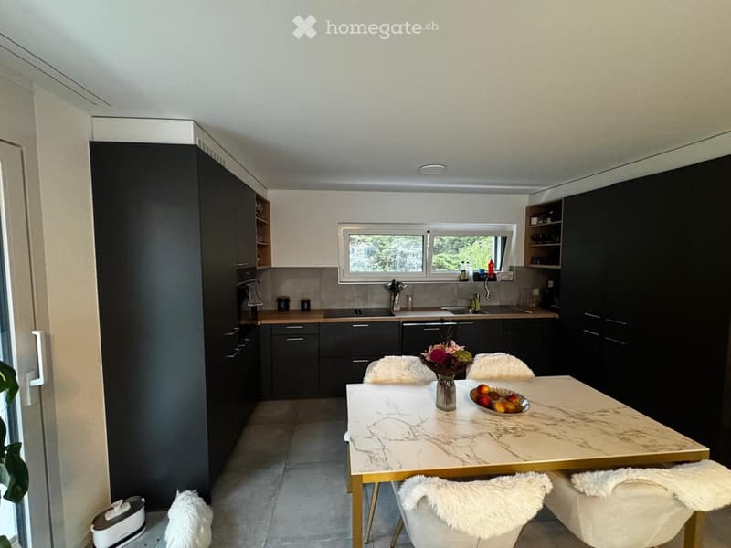 Modern kitchen with dark cabinets, a marble dining table, and four white chairs with fur cushions. The kitchen features a window, a sink, and a dishwasher.