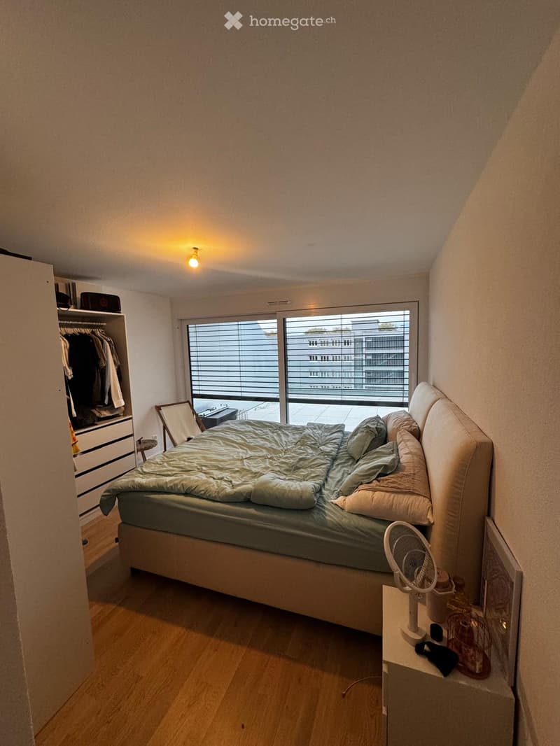 Bedroom with a large bed, nightstand, fan, wardrobe, and a mirror. The room has wooden flooring and large windows with blinds.