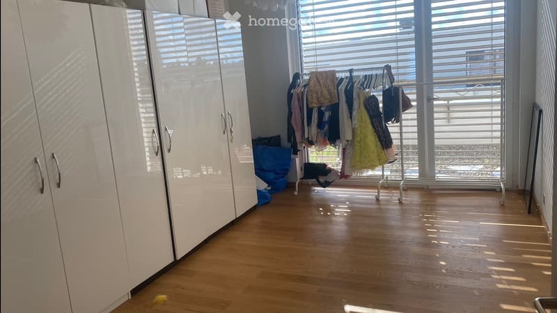 Empty room with wooden flooring, white built-in wardrobes, and a clothes drying rack near the window. The room is well-lit with natural light coming through large windows with blinds.