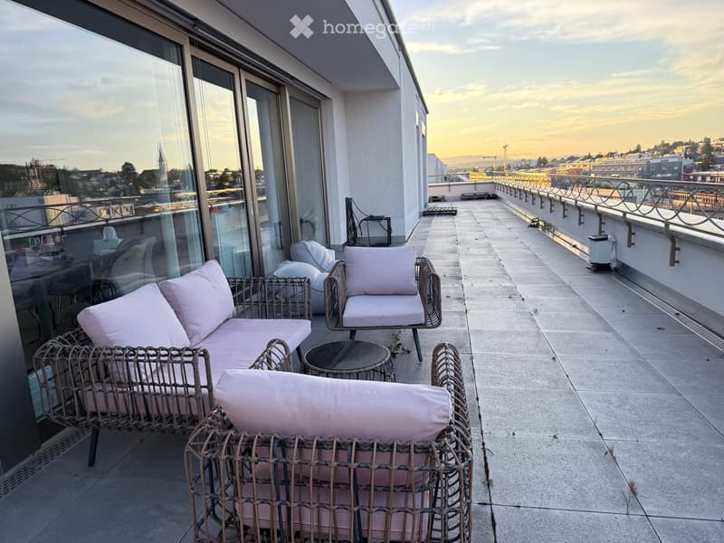 Balcony with wicker furniture, including a sofa, chairs, and a small table, overlooking a cityscape at sunset.