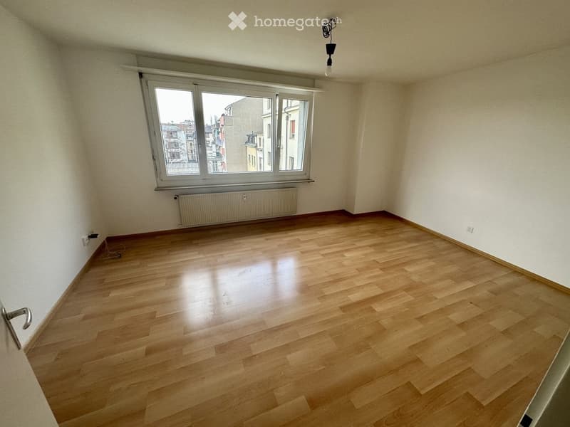 Empty room with wooden flooring, white walls, large window, radiator, and a single light fixture.