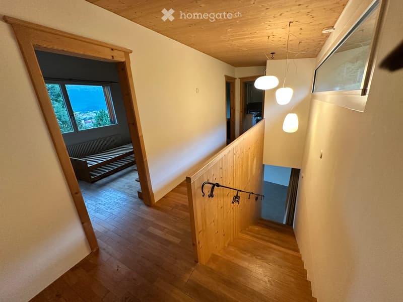 Wooden staircase with a railing, pendant lights, and a view of a room with a bench and large window.