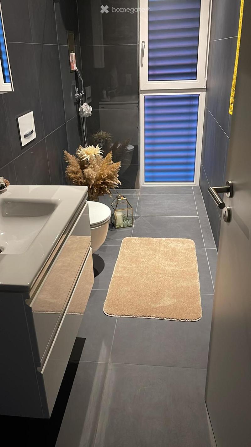 Modern bathroom with dark grey tiles, a white sink, toilet, shower area, and decorative elements including a flower arrangement and a candle holder.