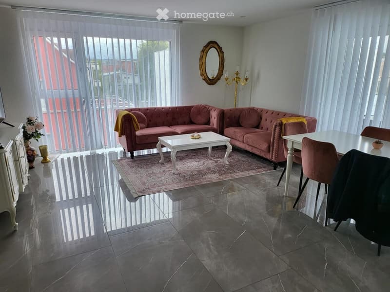 Elegant living room with a pink velvet sofa, white coffee table, dining area with a white table and chairs, large windows with vertical blinds, a patterned area rug, and decorative items including a mirror and candelabra.