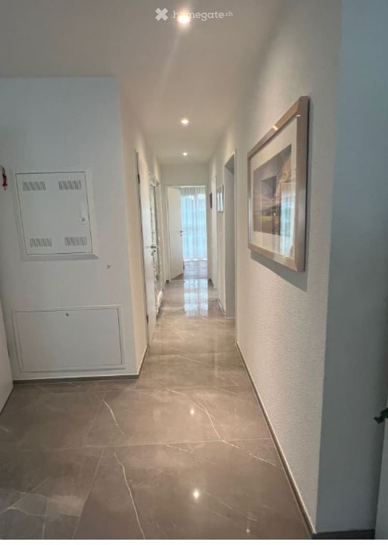 A clean, well-lit hallway with tiled flooring and white walls. There are several doors along the sides, a framed picture on one wall, and a small electrical panel on the left side.