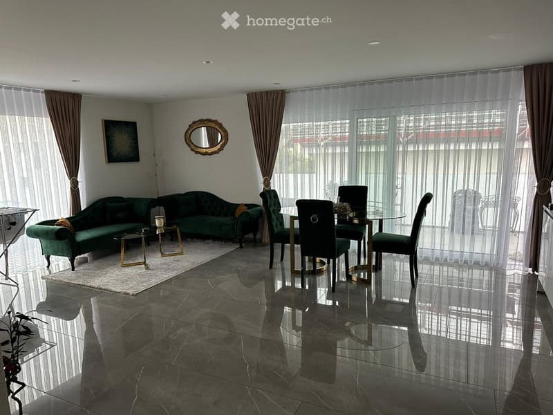 A modern living room with green sofas, a glass dining table, and chairs, large windows with sheer curtains, and a polished tile floor.