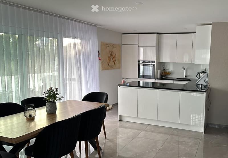 Modern kitchen with white cabinets, black countertops, and a dining area featuring a wooden table and black chairs. A potted plant and candle are on the table, and a poster is on the wall.