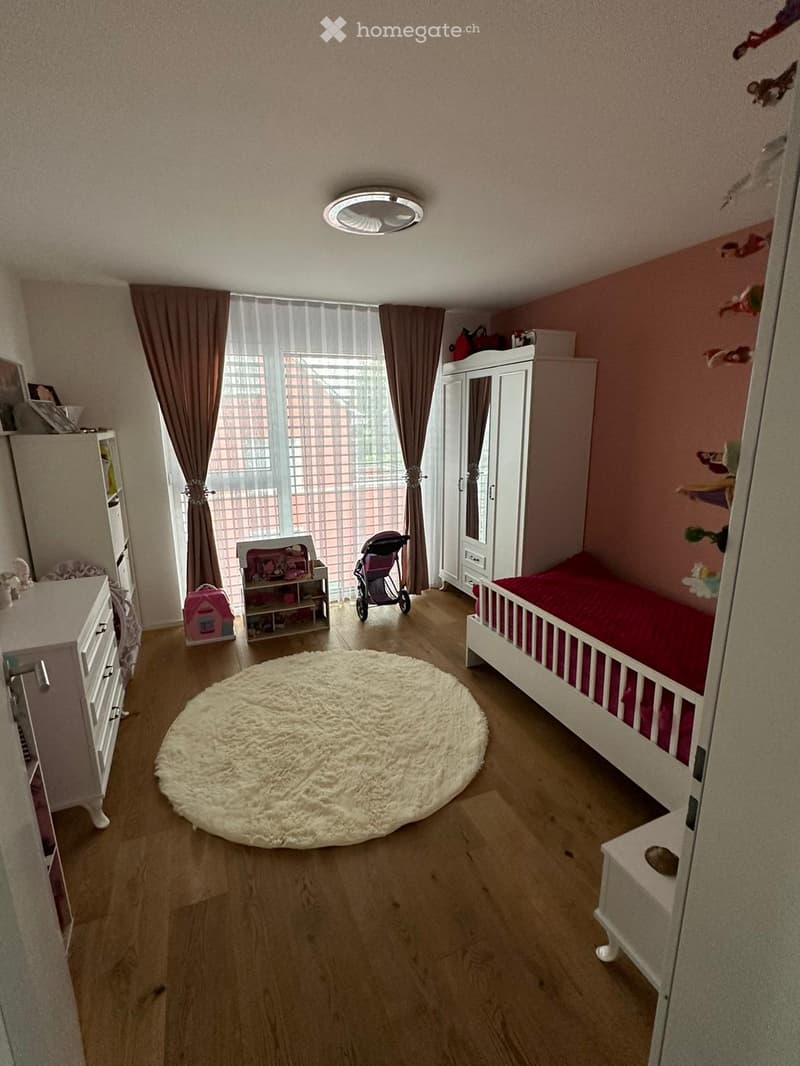 A well-lit children's bedroom with a single bed, wardrobe, drawer unit, round rug, and various toys including a baby stroller and a toy house. The room features wooden flooring and large windows with curtains.