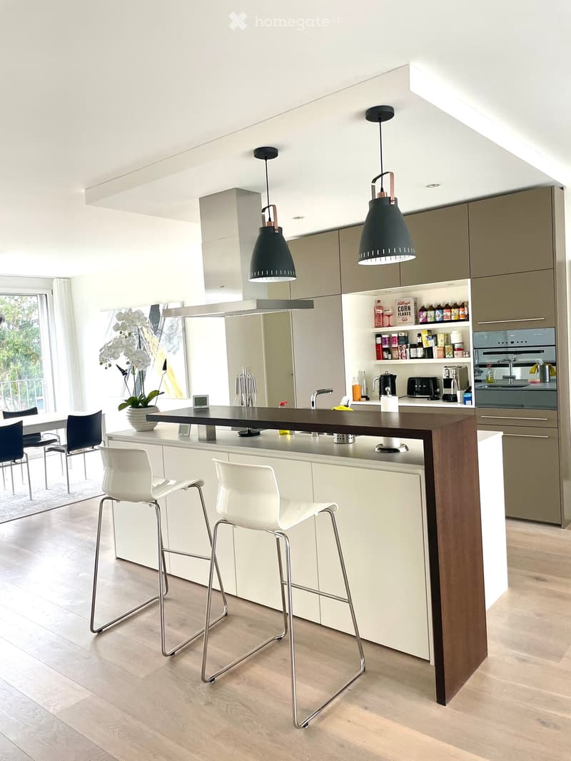 Modern kitchen with a central island, bar stools, and built-in appliances, including an oven and a sink with a faucet. The space features light wood flooring and white cabinets.