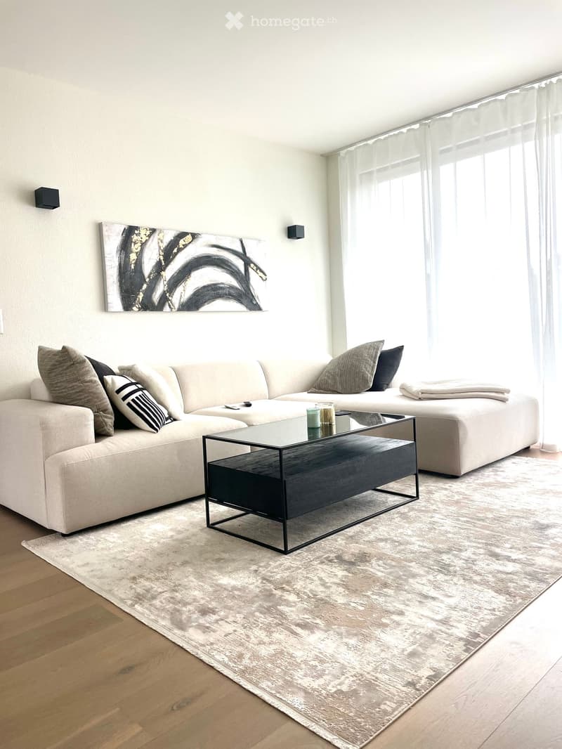 Modern living room with a light-colored sectional sofa, multiple pillows, a sleek black coffee table, and a large abstract artwork on the wall. The room features wooden flooring, a large area rug, and large windows with sheer curtains.