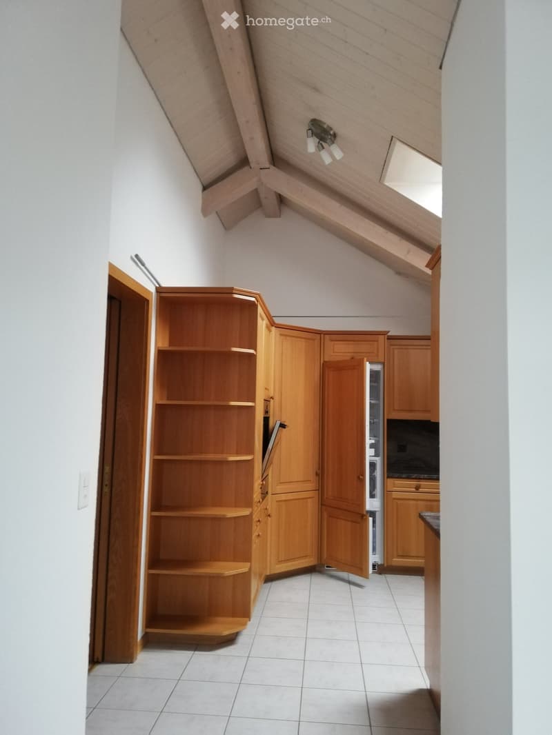 A small kitchen with wooden cabinets, a refrigerator, and a shelf unit. The ceiling has exposed wooden beams and a skylight.