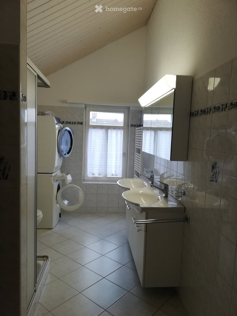 Modern bathroom with double sink, mirror, washing machine, toilet, and large window letting in natural light.