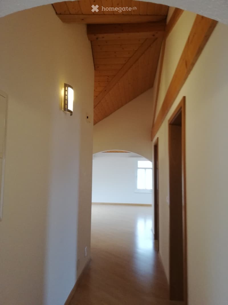 A narrow hallway with wooden beams on the ceiling, white walls, and a small window at the end. The floor is polished wood, and there is a wall-mounted light fixture on the left side.