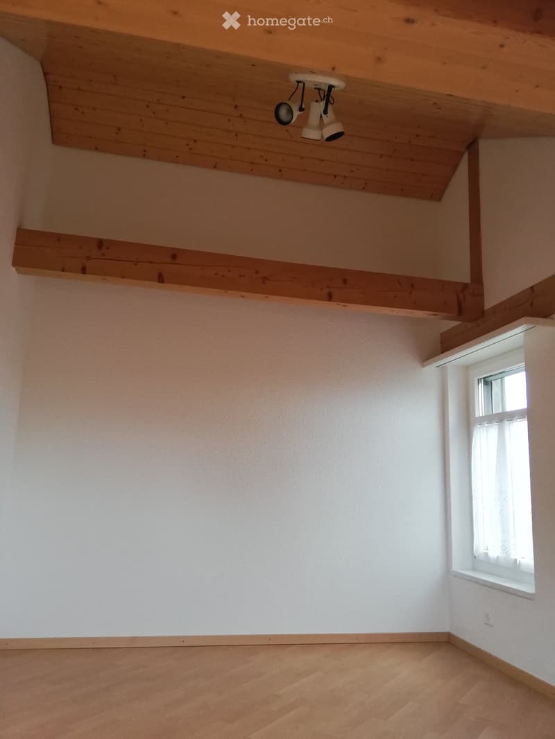 Empty room with wooden floor, white walls, and a small window with white frame and curtain. Exposed wooden ceiling beams and a built-in shelf.