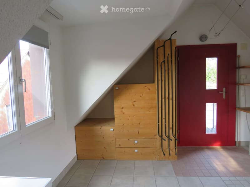 An attic room with slanted ceilings, tiled floor, large window, wooden built-in storage unit, and a red door.
