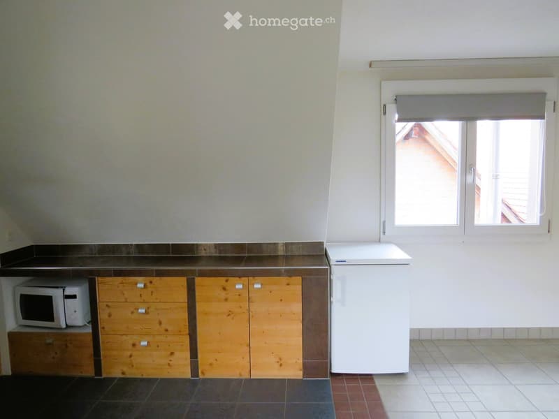 Kitchen with wooden cabinets, a microwave, and a refrigerator. The space has tiled flooring and a window with blinds.