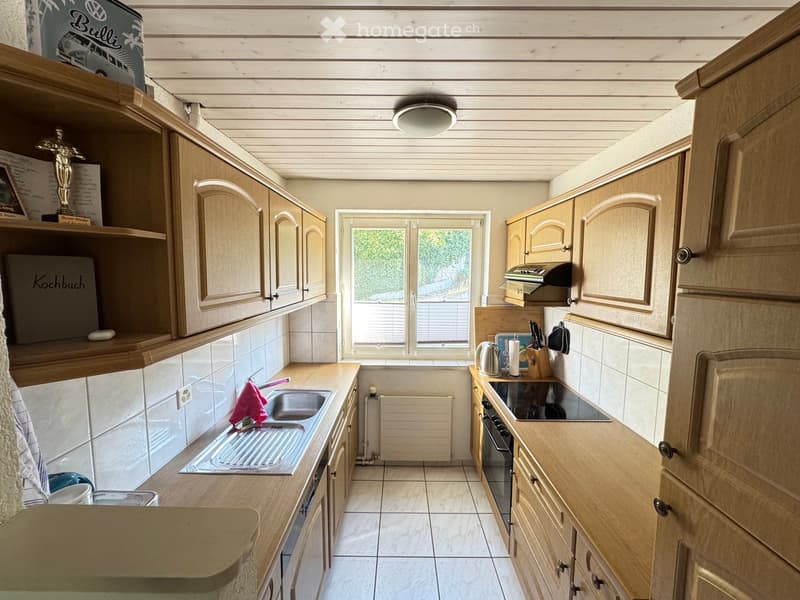 The kitchen features light wooden cabinets, a central island, and a sink with a faucet. Appliances include an oven and stove, and there are various kitchen items such as a cup, knife block, and a radiator under the window.