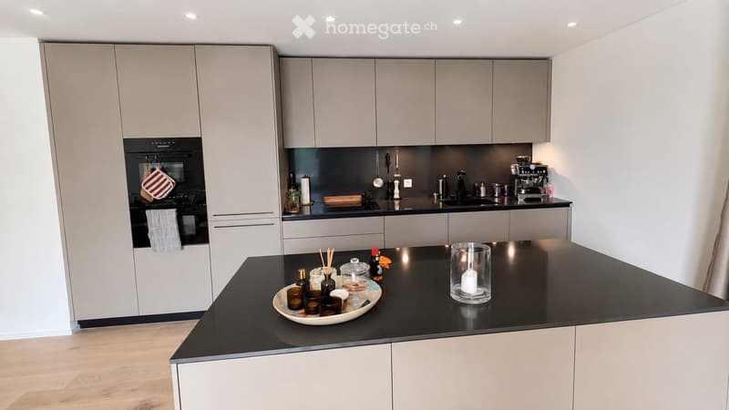 Modern kitchen with light grey cabinets, black countertops, and various appliances including an oven, coffee maker, and sink. A candle and decorative items are placed on the island.