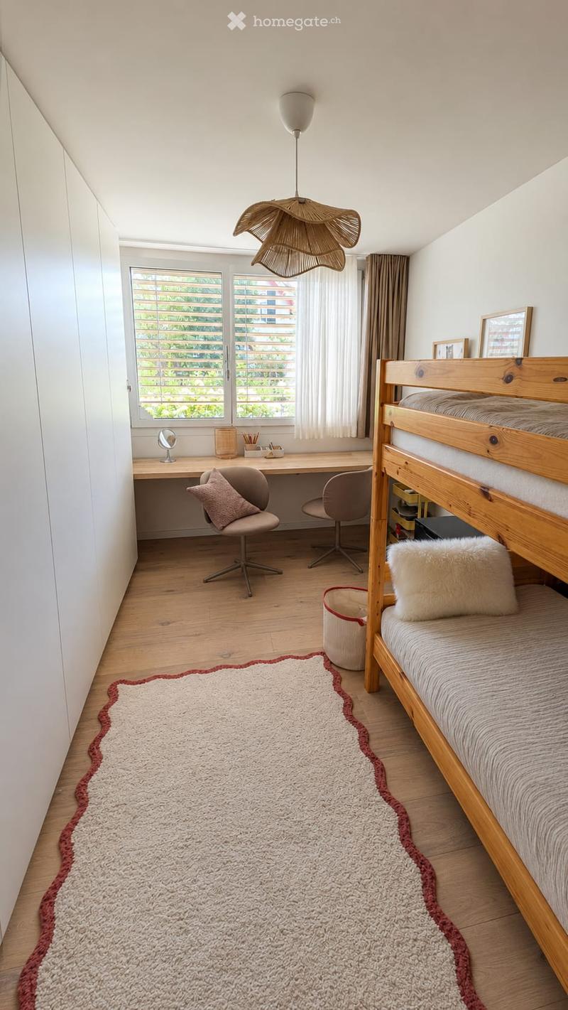 A modern bedroom with a wooden bunk bed, a desk with a chair, a mirror, and a large window with blinds. The room features a cozy rug with a red border and a stylish hanging lamp.