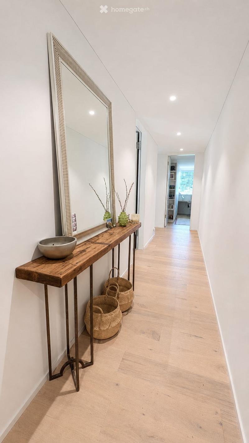 Modern hallway with light wooden flooring, white walls, and a long wooden console table with a mirror, decorative items, and woven baskets.
