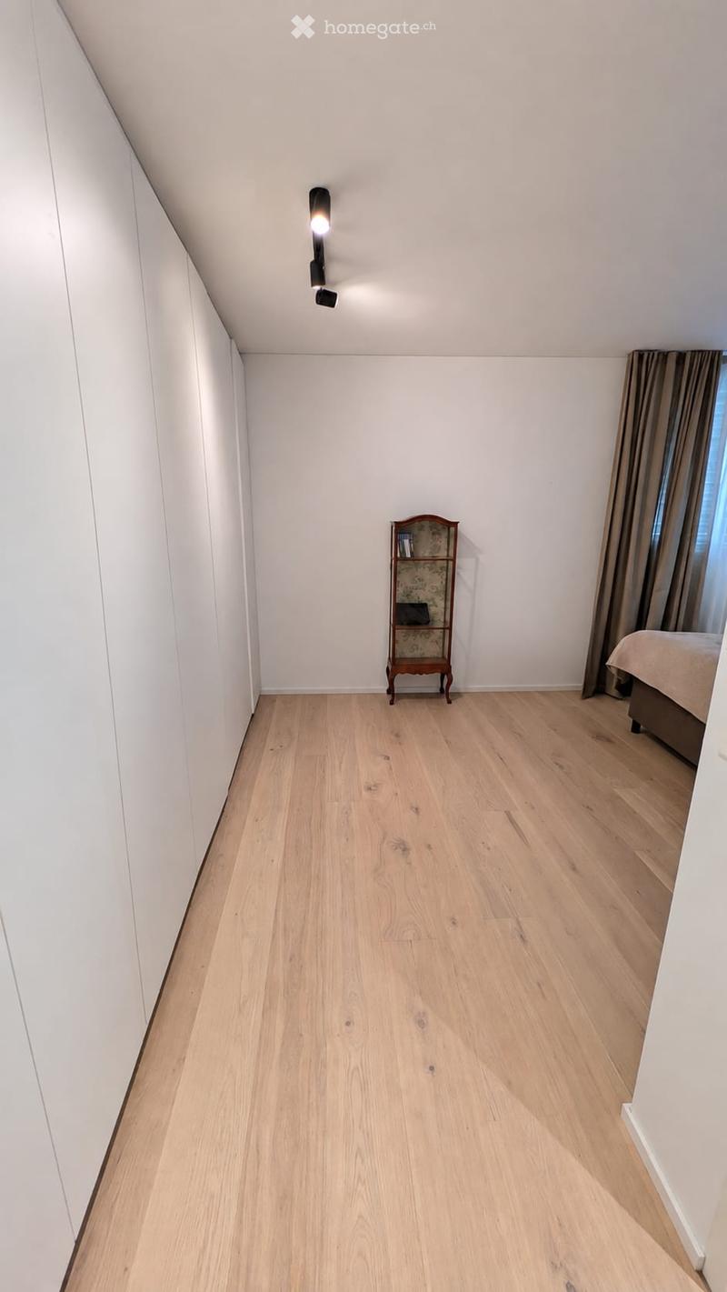 A small, modern bedroom with light wooden flooring, white walls, and a slanted ceiling. Features a bed with a beige duvet, a small wooden nightstand with a decorative patterned design, and a curtained window.