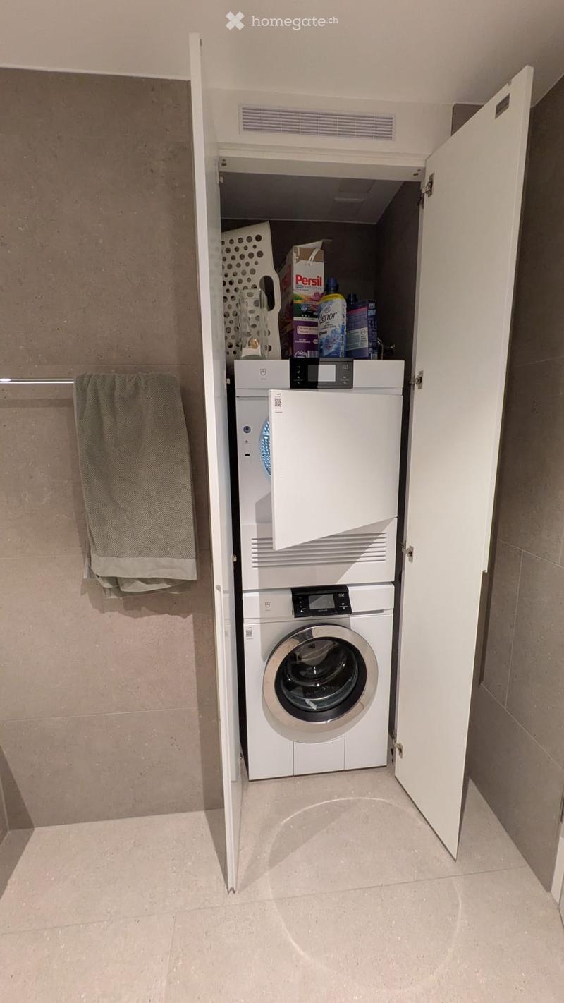 Compact bathroom with a stacked washing machine and dishwasher unit, a towel hanging on a rack, and cleaning supplies stored on top of the appliances.
