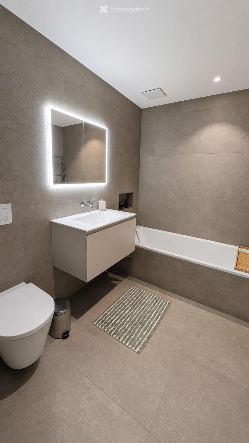 Modern bathroom with a sleek sink, illuminated mirror, bathtub, toilet, and a small trash can. The floor is tiled, and a striped bath mat is placed in front of the sink.