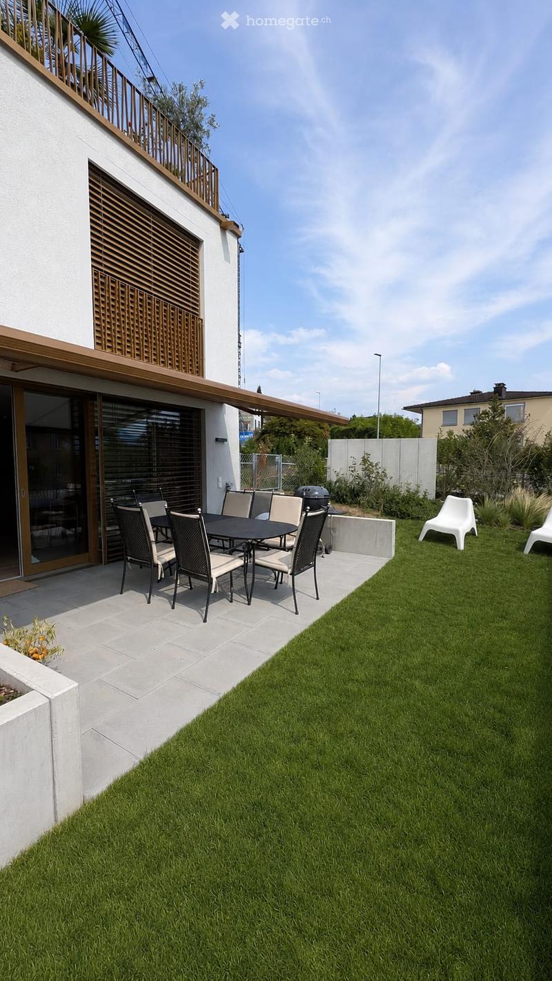 Garden with a well-maintained lawn, patio area with a dining table and chairs, a grill, and two white lounge chairs.