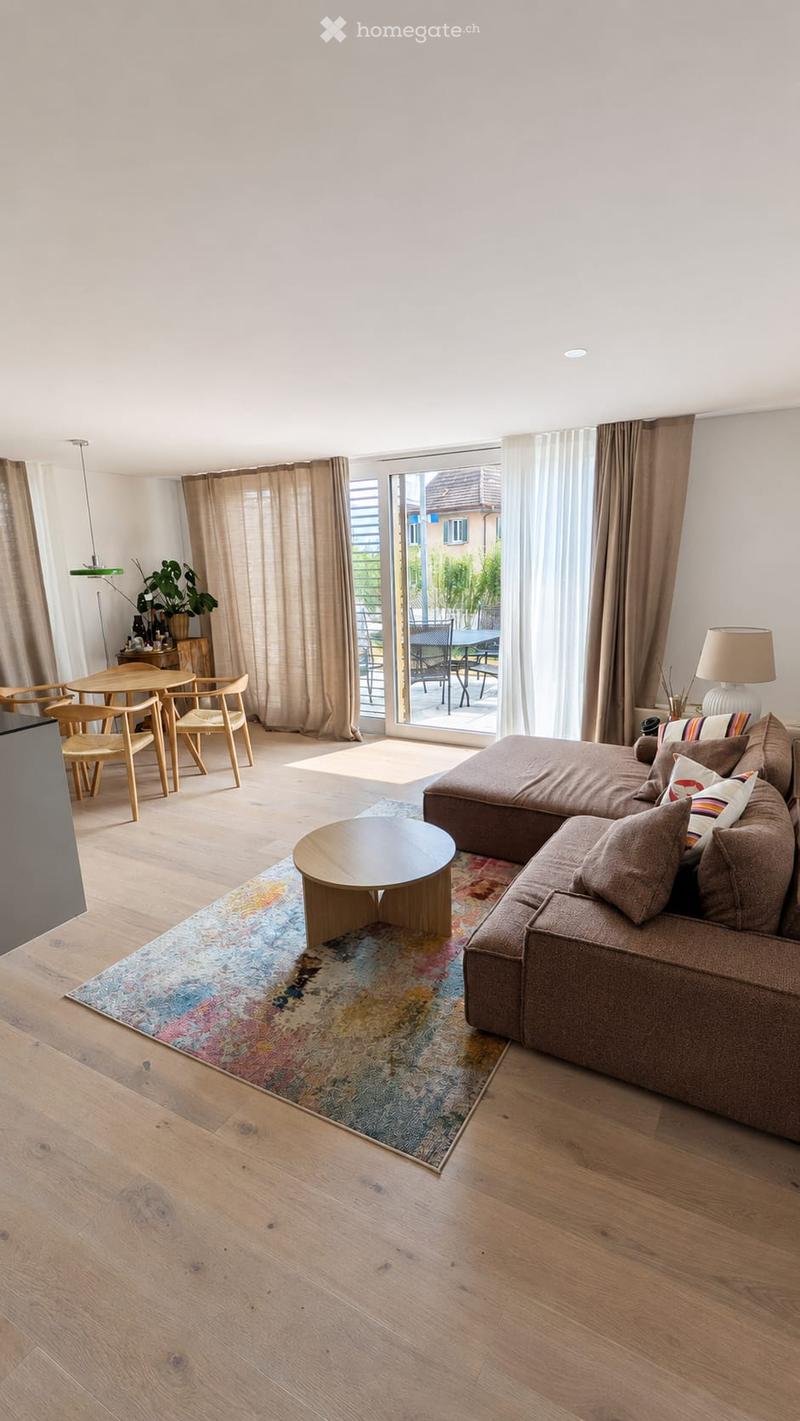 Modern living room with wooden flooring, a large sectional sofa, coffee table, dining area with chairs, and large windows leading to a balcony.