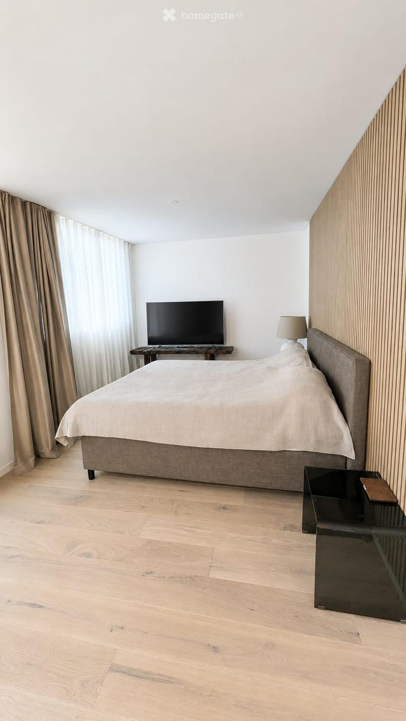 Modern bedroom with a large bed, nightstand, table lamp, television on a wooden stand, and a side table with a book.