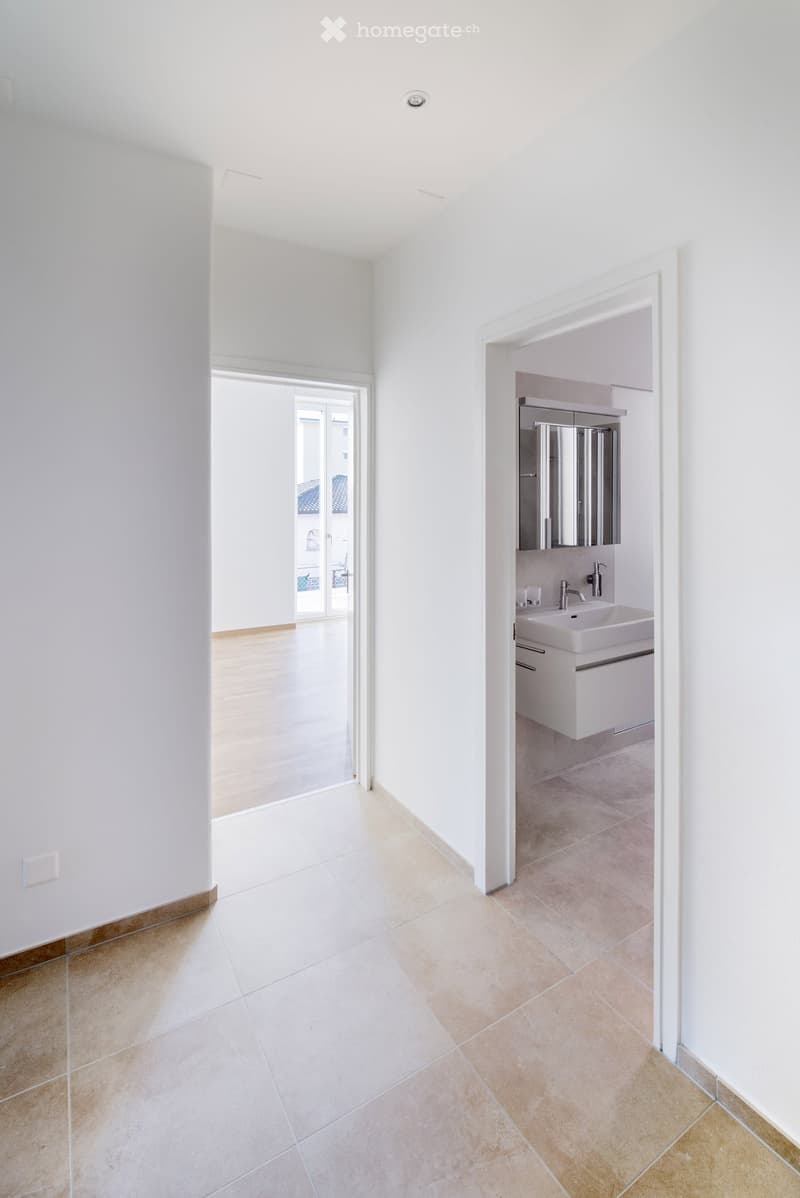 Bright, minimalist hallway with tiled flooring and white walls, leading to a modern bathroom with a sink, mirror, and cabinet.
