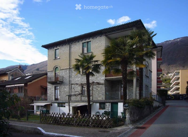 A multi-story stone building with balconies and palm trees in front, located in a residential area with a clear sky and distant mountains.