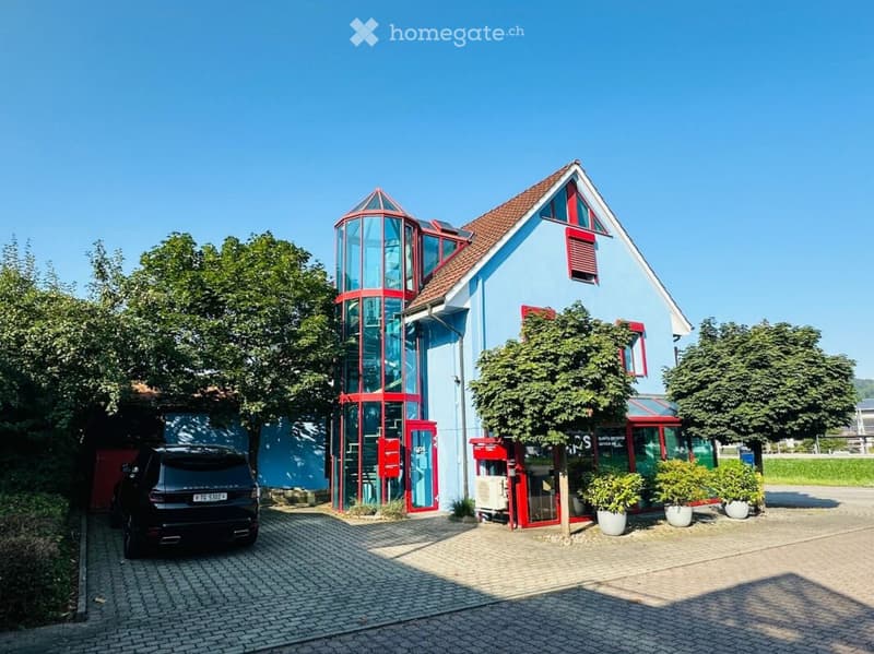 Modern two-story building with light blue walls, red accents, and a red cylindrical tower. Features a paved driveway, parked car, and small garden area with potted plants.