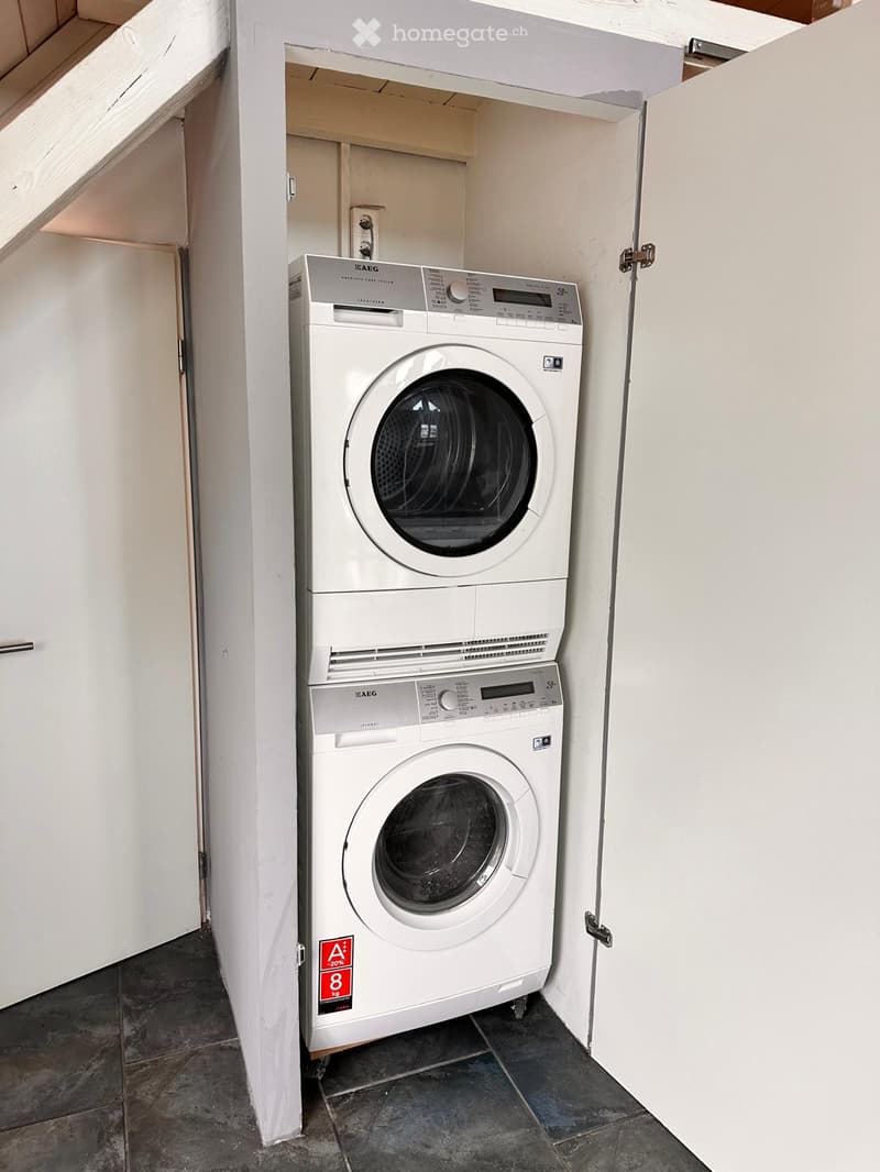 Compact bathroom with a stacked washing machine and dishwasher unit, white tiled walls, and dark tiled floor.