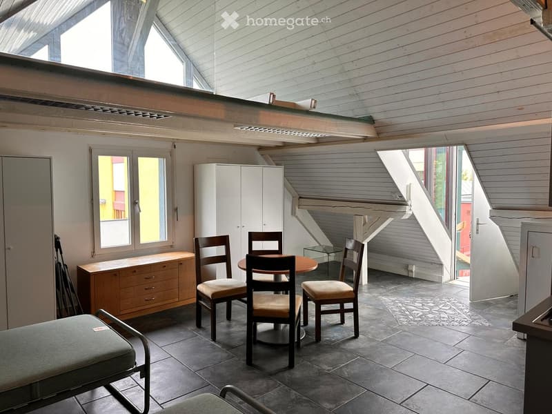 Bright, airy room with exposed wooden beams and skylights, featuring a round wooden table with four chairs, a bench, and a tall white cabinet.