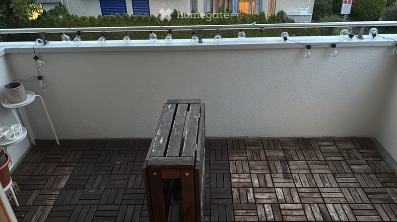 Balcony with wooden decking, a bench, small table with cups, string lights, and potted plants.