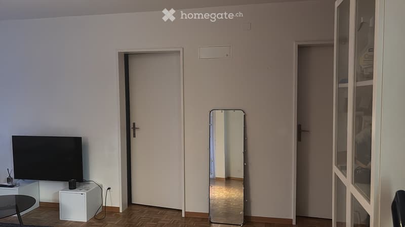 A modern, minimalistic living room with a television on a white stand, a black chair, a small table, and a large mirror reflecting part of the room.