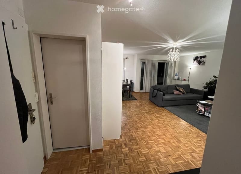 Open-plan living and dining area with wooden herringbone flooring, grey sofa, coffee table, dining table with chairs, and a hanging light fixture.