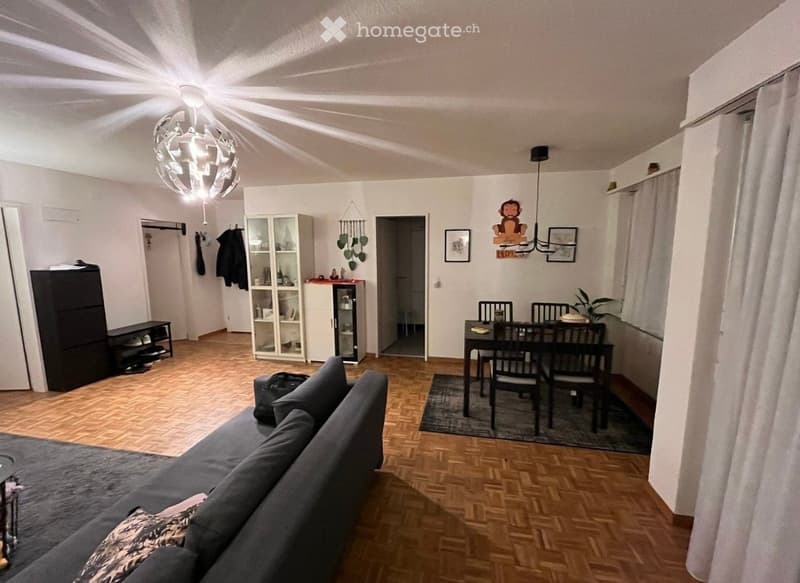 Open-plan living and dining area with wooden flooring, a grey sofa, a dining table with chairs, a cabinet, a potted plant, and a chandelier.