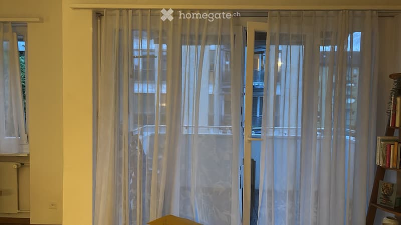 View of a room with large windows and sheer curtains, leading to a balcony with a view of the city. A small shelf with books is visible on the right side.