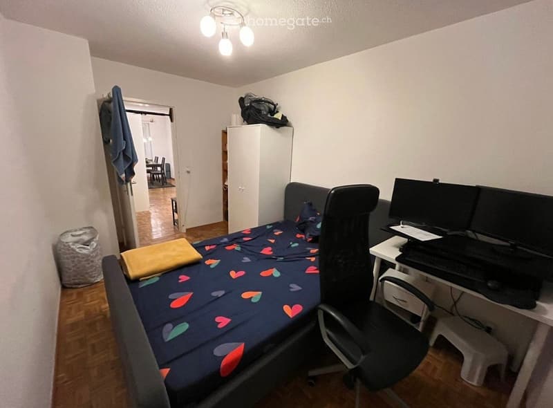 A furnished bedroom with a bed, desk, chair, and wardrobe. The room features wooden flooring and white walls.