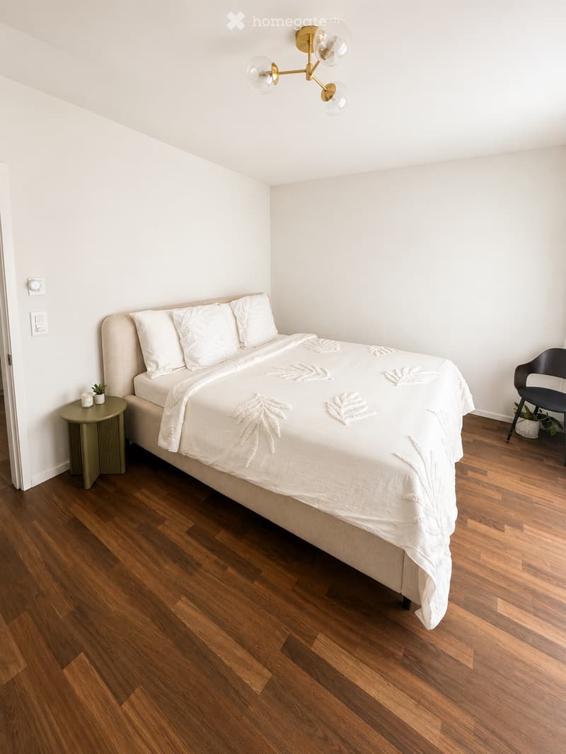 A modern bedroom with a large bed, white bedding, a nightstand with a lamp, a small round table, and a black chair. The room features wooden flooring and white walls.