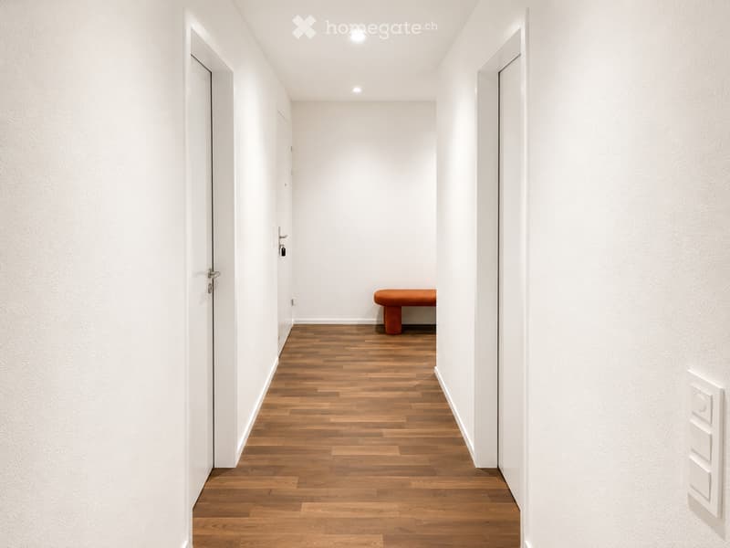 A narrow hallway with wooden flooring, white walls, and a small orange stool placed against the wall.