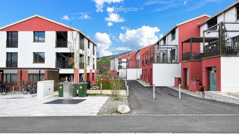 Modern residential complex with red and white facades, multiple balconies, and a paved courtyard with bicycles and trash bins.