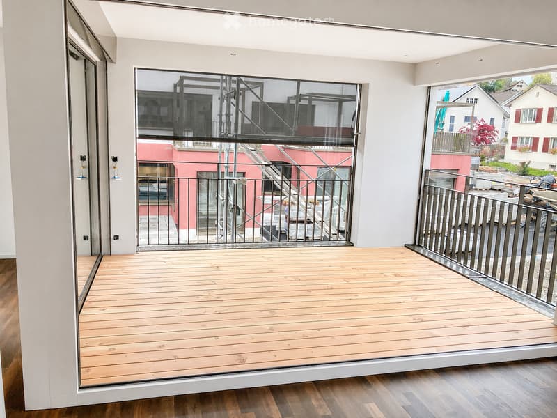 Modern wooden balcony with glass railings, overlooking a cityscape with residential buildings and greenery.