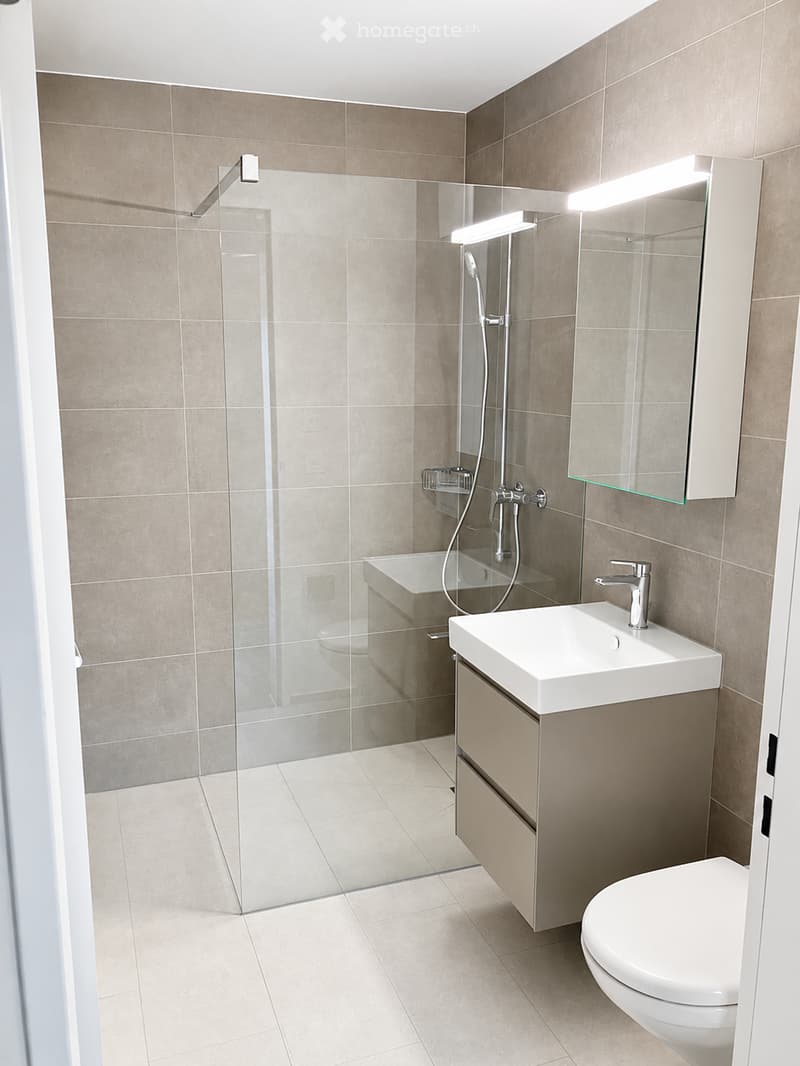 Modern bathroom with beige tiles, a white sink with a chrome faucet, a large mirror with built-in lighting, a glass-enclosed shower, and a toilet.