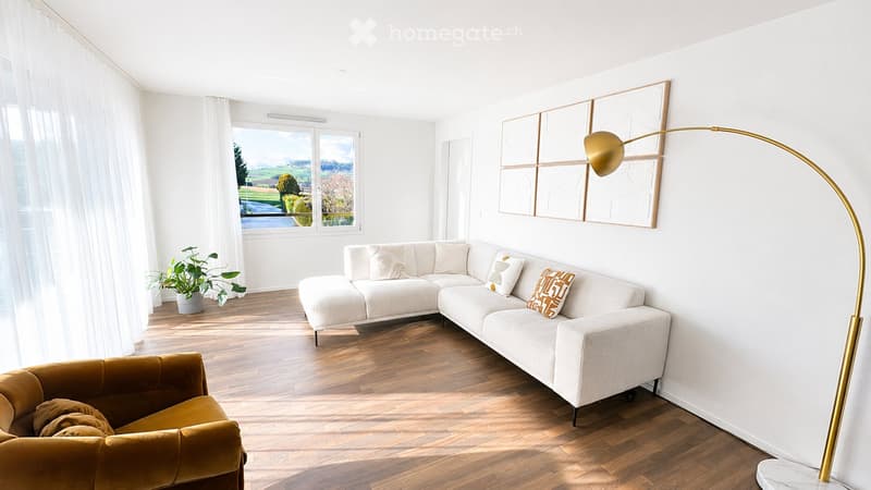 Modern living room with white walls, wooden flooring, large windows, a white sectional sofa, a mustard-colored armchair, a floor lamp, a potted plant, and artwork on the wall.