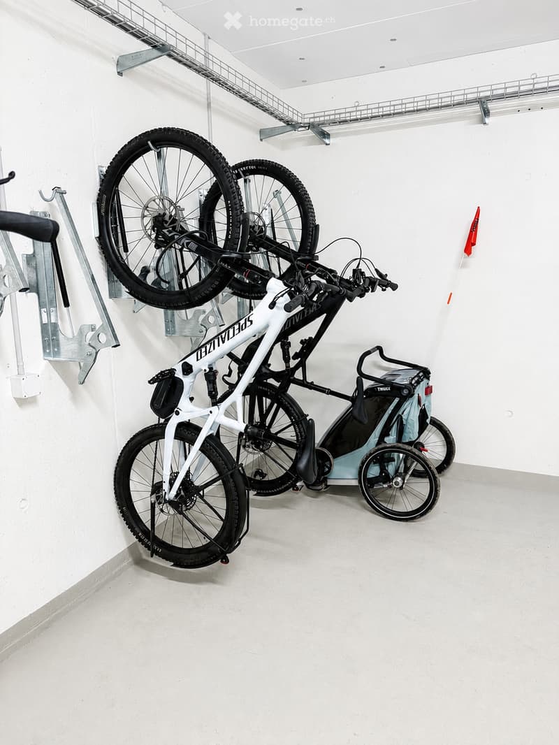 A clean, modern storage area with bicycles and a baby stroller neatly arranged. The space features white walls, a concrete floor, and a metal ceiling with integrated lighting.