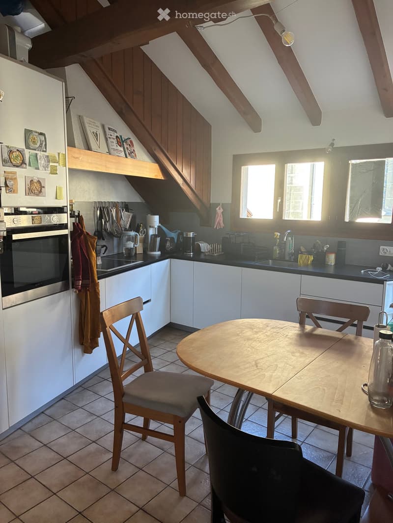 Bright kitchen with white cabinets, stainless steel appliances, and a wooden dining table with chairs. The space features a tiled floor and large windows allowing natural light to fill the room.