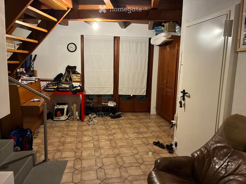 A cozy hallway with a wooden staircase, a red table with shoes and bags, a pet carrier, and a brown leather sofa on the right side.